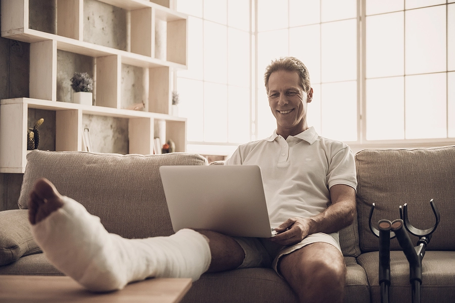 Middle aged man with a cast on his leg sitting on his couch searching for the best injury lawyer